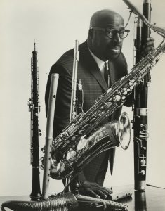 black & white photograph of Yusef Lateef posing with some of his instruments, including an oboe, flute, and baritone saxophone. Lateef is wearing a suit and tie, glasses, and he has a mustache and short goatee